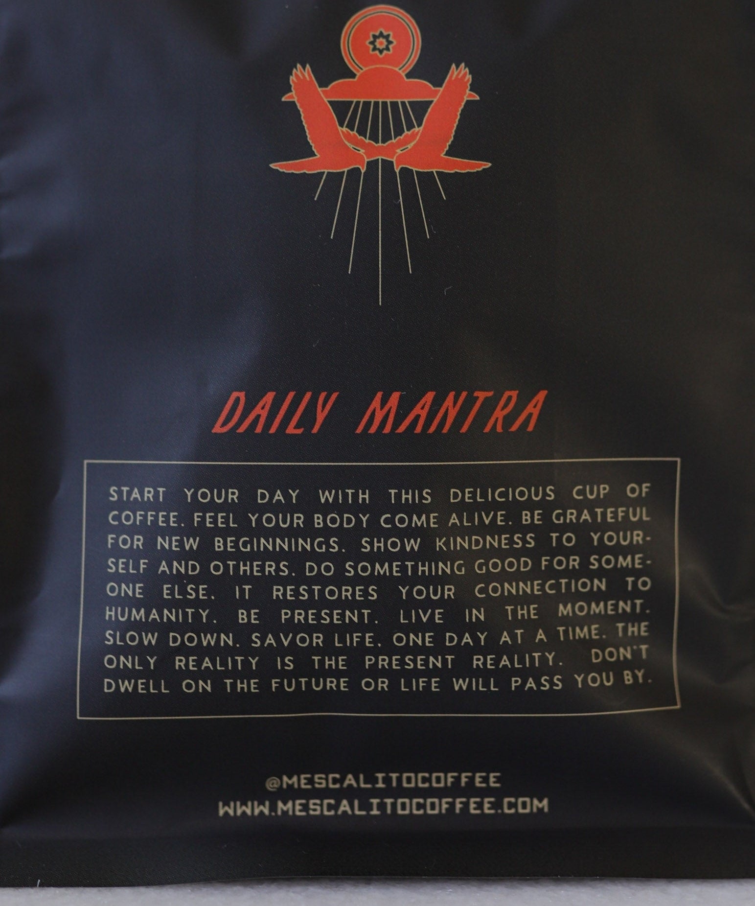 Black coffee bag labeled "Desert Bloom" by Mescalito Coffee, Whole Blend beans. Features motivational text inspiring mindfulness, kindness, and presence. Roasted in Brenham, TX. Website "mescalitocoffee.com" appears at the bottom.