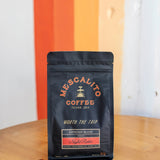 A black bag of Mescalito Coffee's Night Rider Blend Whole Coffee Beans sits on a wooden table in front of a wall with orange, white, and beige vertical stripes.