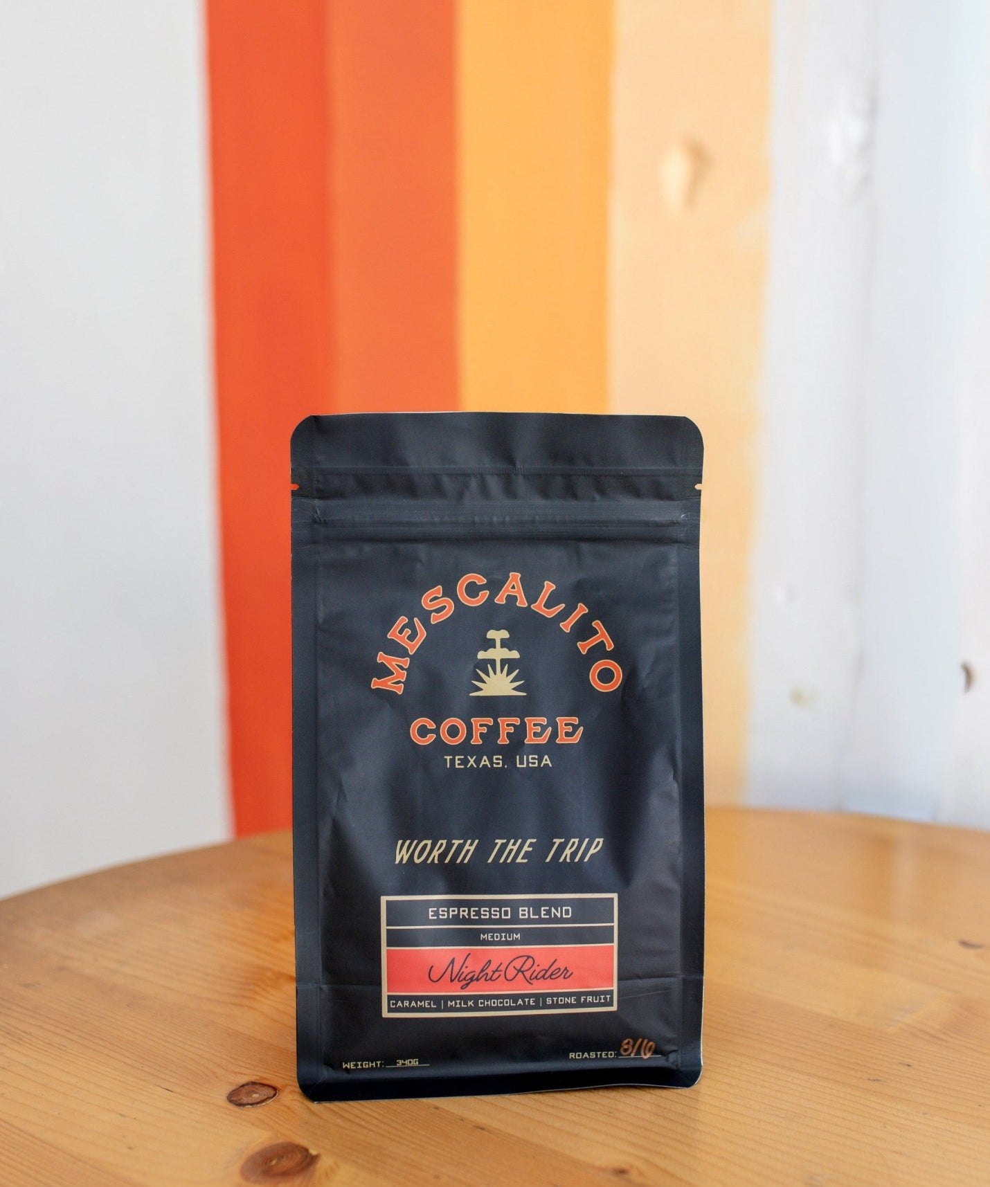 A black bag of Mescalito Coffee's Night Rider Blend Whole Coffee Beans sits on a wooden table in front of a wall with orange, white, and beige vertical stripes.