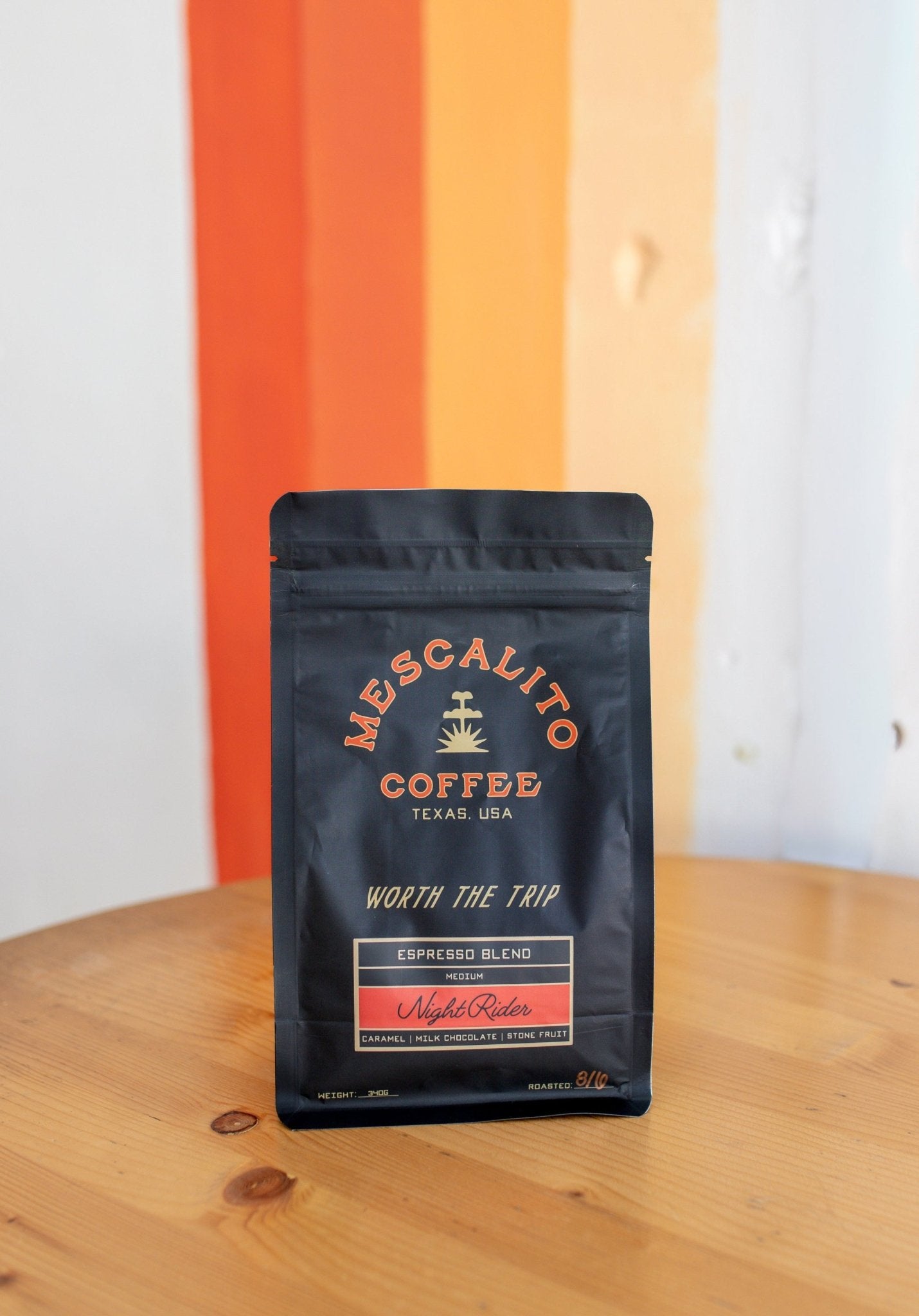 A black bag of Mescalito Coffee's Night Rider Blend Whole Coffee Beans sits on a wooden table in front of a wall with orange, white, and beige vertical stripes.