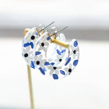 Blue Acrylic Flower Hoops by Jill Makes feature hand-painted blue and white floral patterns on clear acrylic. Lead and nickel free for comfortable wear, these unique earrings are displayed on a gold stand against a blurred background.