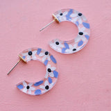 Blue Acrylic Flower Hoops by Jill Makes are clear, crescent-shaped earrings featuring blue, white, and black floral designs. These hand-painted, lead and nickel free earrings artistically elevate any outfit on a pink textured surface.