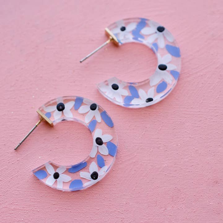 Blue Acrylic Flower Hoops by Jill Makes are clear, crescent-shaped earrings featuring blue, white, and black floral designs. These hand-painted, lead and nickel free earrings artistically elevate any outfit on a pink textured surface.
