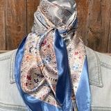 The Blue Floral Wild Rag by Rodeo Drive, featuring shiny silk with blue and cream floral patterns, is tied around the neck of someone in a light denim jacket against a wooden background.