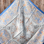 The Blue Floral Wild Rag by Rodeo Drive, featuring intricate blue, orange, and grey paisley and floral patterns, is displayed spread out on a wooden surface.
