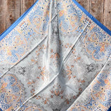 The Blue Floral Wild Rag by Rodeo Drive, featuring intricate blue, orange, and grey paisley and floral patterns, is displayed spread out on a wooden surface.