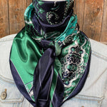 A person wears the Rodeo Drive Blue & Green Waves Wild Rag tied around their neck over a light denim jacket, set against a wooden background.