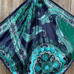 The Blue & Green Waves Wild Rag by Rodeo Drive features an ocean-inspired paisley pattern in blue, teal, and green, elegantly displayed against a wooden background.