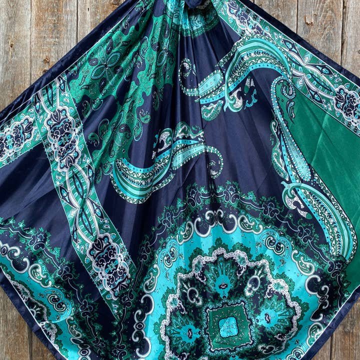 The Blue & Green Waves Wild Rag by Rodeo Drive features an ocean-inspired paisley pattern in blue, teal, and green, elegantly displayed against a wooden background.
