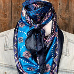 A person wears the Rodeo Drive Blue & Purple Paisley Wild Rag scarf tied around their neck, styled over a light denim jacket against a wooden background.