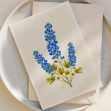 The Bluebonnets Trio Blank Greeting Card by Annie Bailey Art, featuring illustrated North American wildflowers, is displayed on a speckled white plate with its matching envelope.
