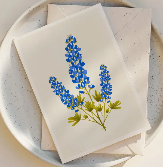 The Bluebonnets Trio Blank Greeting Card by Annie Bailey Art, featuring illustrated North American wildflowers, is displayed on a speckled white plate with its matching envelope.