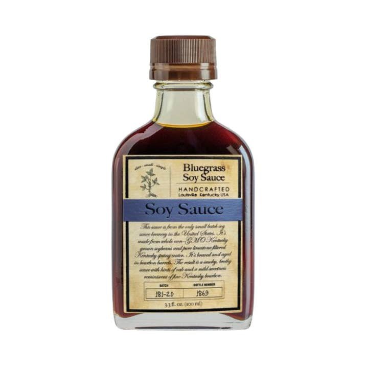A glass bottle of Bluegrass Soy Sauce Bourbon Barrel Foods by Bourbon Barrel Foods features a brown cap, a blue-accented label, and delivers authentic Kentucky soy sauce with bourbon barrel-aged richness.