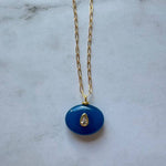 The Boheme Gemstone Pendant Necklace by Jessica Matrasko Jewelry features a gold filled chain with a blue oval pendant and teardrop-shaped clear stone. Personalize it by adding customizable charms. Displayed on a light gray surface.