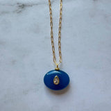 The Boheme Gemstone Pendant Necklace by Jessica Matrasko Jewelry features a gold filled chain with a blue oval pendant and teardrop-shaped clear stone. Personalize it by adding customizable charms. Displayed on a light gray surface.