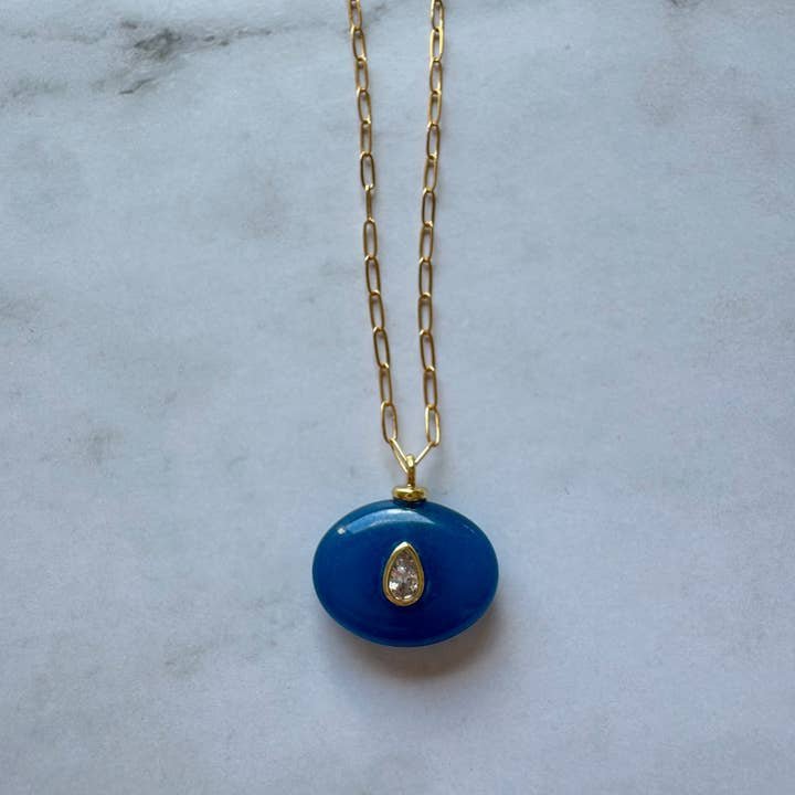 The Boheme Gemstone Pendant Necklace by Jessica Matrasko Jewelry features a gold filled chain with a blue oval pendant and teardrop-shaped clear stone. Personalize it by adding customizable charms. Displayed on a light gray surface.