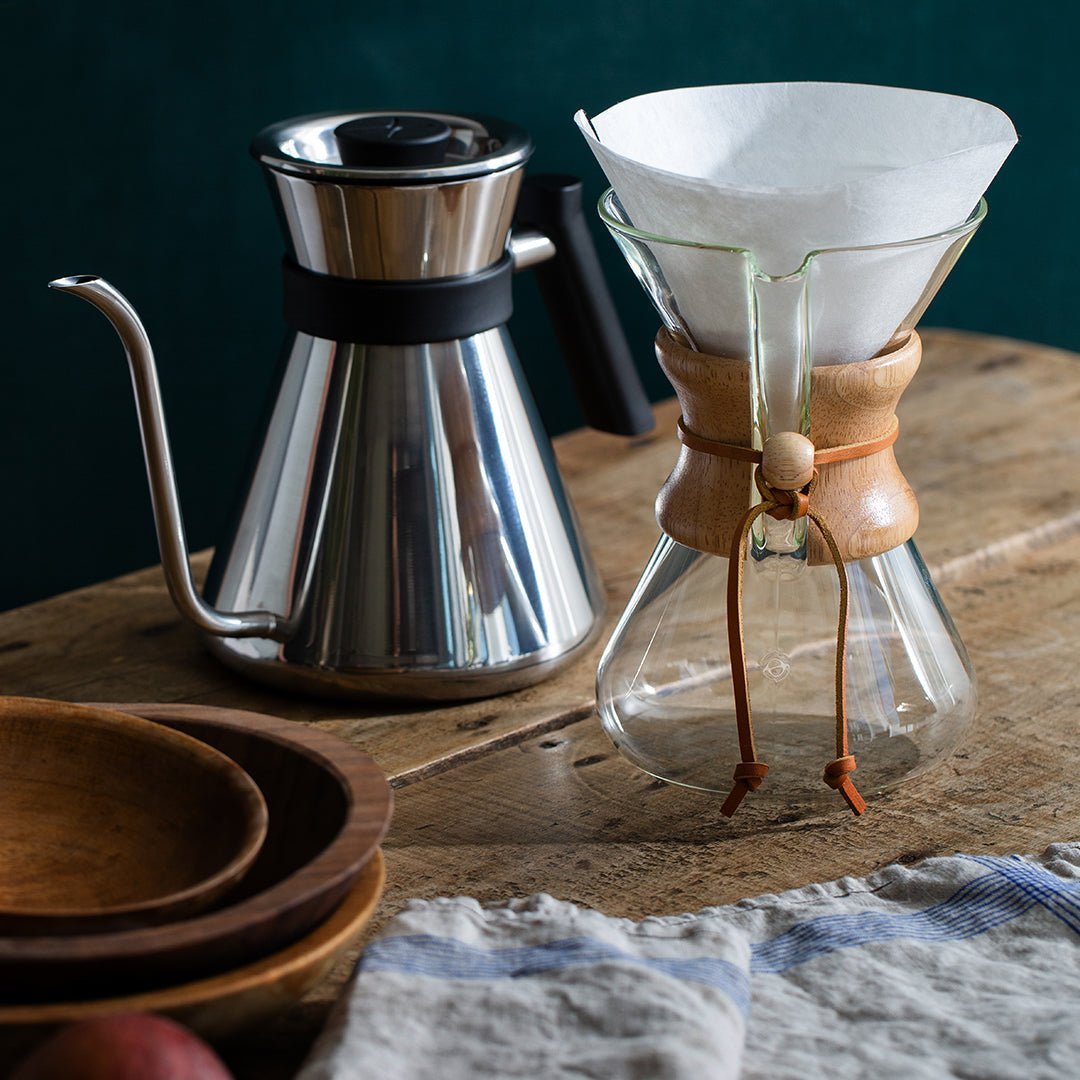 A stainless steel gooseneck kettle and a glass Chemex coffee maker with Chemex Bonded Filters | Circle sit on a wooden table beside wooden bowls and a cloth napkin, perfect for pourover brewing.