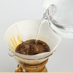 A glass kettle pours hot water over ground coffee in a paper filter inside a Chemex, demonstrating classic pourover brewing with Chemex Bonded Filters | Circle by Chemex.
