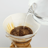A glass kettle pours hot water over ground coffee in a paper filter inside a Chemex, demonstrating classic pourover brewing with Chemex Bonded Filters | Circle by Chemex.