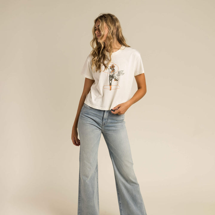A woman wears the Boot Scootin' Boxy Tee by Sendero Provisions Co., paired with light blue high-waisted wide-leg jeans and cowboy boots, standing against a plain beige background for a Western-inspired look.