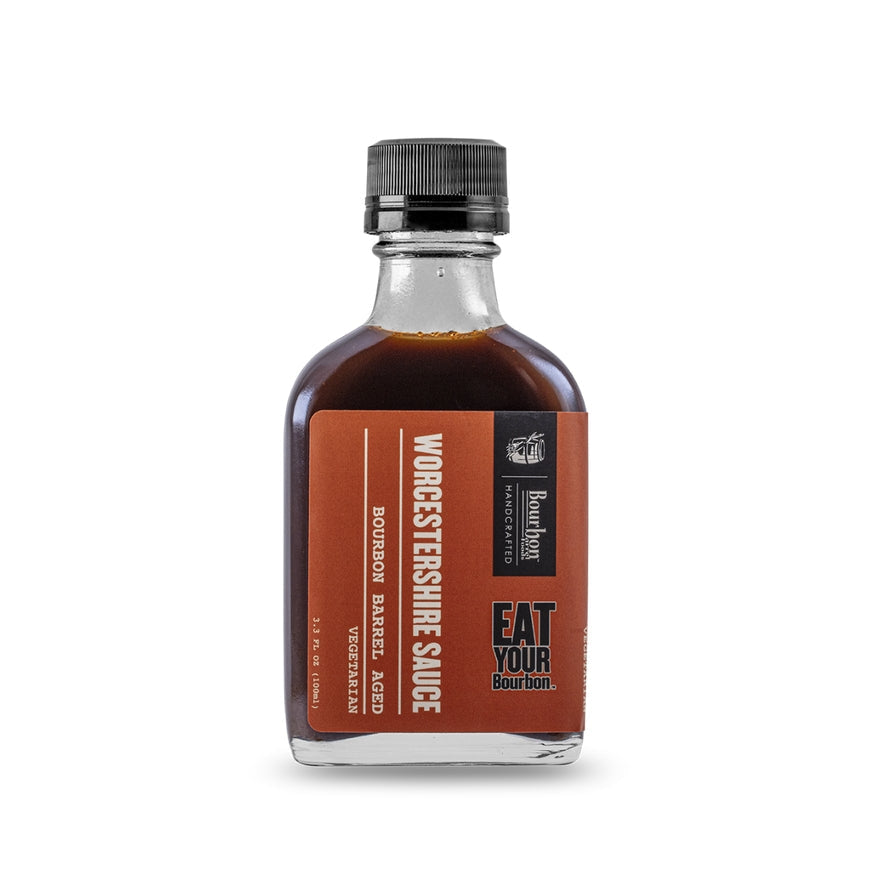 A clear glass bottle with a black cap, labeled "Bourbon Barrel Aged Worcestershire" by Bourbon Barrel Foods. This all-natural, vegetarian sauce features the “Eat Your Bourbon” branding on the label.