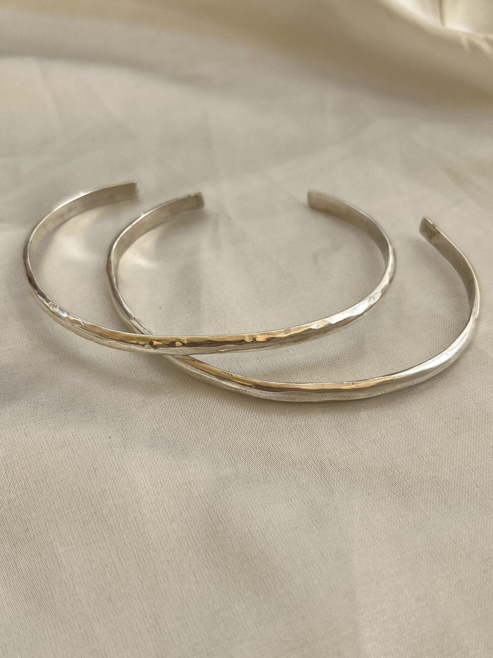 The Simple Silver Hammered Bracelet by Bird of Prey, a small handcrafted piece, is displayed on a light beige fabric surface.