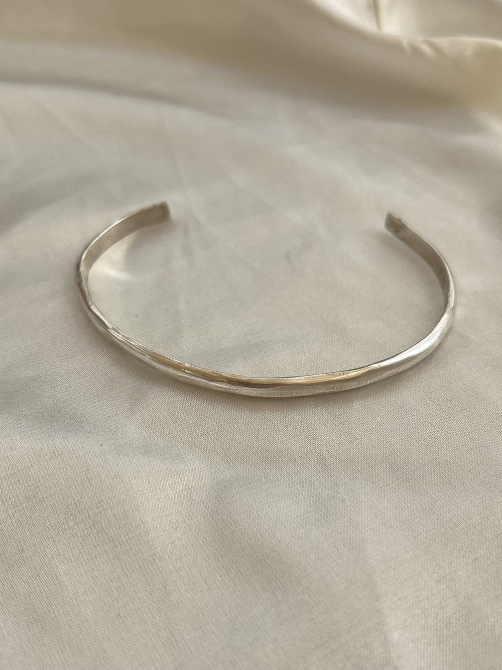 The Bird of Prey Simple Silver Hammered Bracelet, a thin handcrafted sterling silver cuff, is displayed on a light-colored fabric surface.