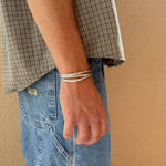 A person in a plaid short-sleeve shirt and blue jeans stands against a beige wall, wearing the Bird of Prey Simple Silver Hammered Bracelet (Small) with their arm relaxed at their side.