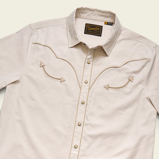 The Broncbuster Snapshirt by Howler Brothers is a beige western shirt with pearl snap buttons and decorative chest stitching, shown laid flat on a light background.