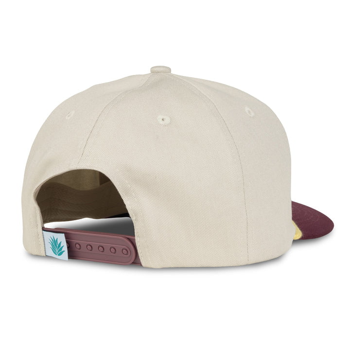 The Buena Suerte Hat by Sendero Provisions Co. is a beige 5-panel cap with a curved visor, seen from the back, featuring a maroon adjustable snap closure and a small tag displaying a blue leaf graphic.