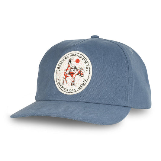 The Burro Lady Hat by Sendero Provisions Co. is a blue cotton twill 5-panel snapback, featuring a circular patch of the Burro Lady riding a horse with "Sendero Provisions Co. Espíritu del Oeste" text.