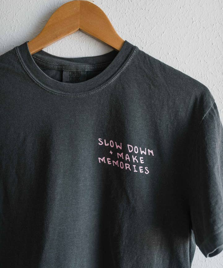 A black “Take the backroads Crop” t-shirt by Ramble & Company hangs on a wooden hanger, featuring the words “SLOW DOWN MAKE MEMORIES” in light pink text on the left chest area. This boxy fit crop top combines style and comfort, making it ideal for everyday wear.
