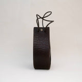 A dark brown leather strap, looped at the top with a string threaded through two holes, stands upright against a plain background—an understated reflection of refined minimalism in the Stash Joi Bag in Coffee.