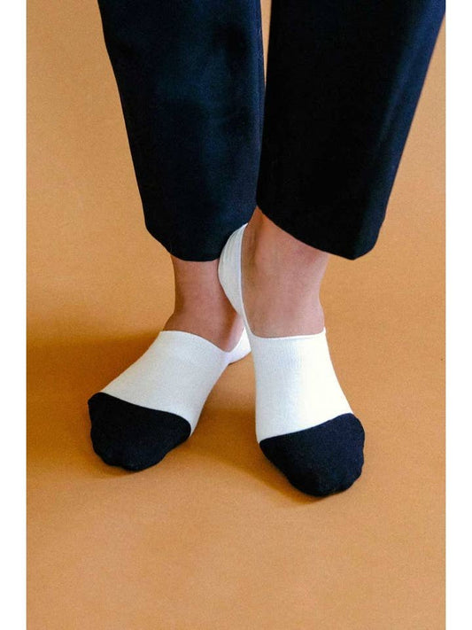 A person wearing Tailored Union Cache 2-Pack Socks, white with black toes and arch support, stands on a light brown floor with feet crossed.