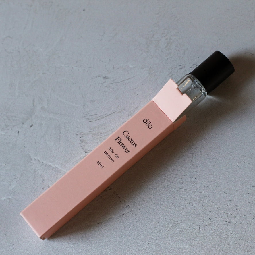 A glass Cactus Flower Eau De Parfum | dilo vial with a black cap sits partly inside its pink box on a light gray surface, hinting at the fresh jasmine notes of this dilo fragrance.
