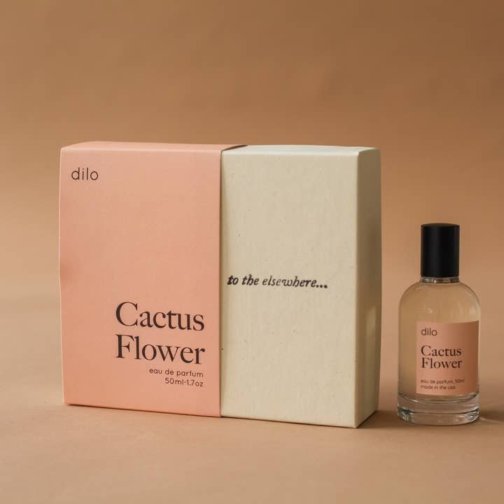 A bottle of dilo's Cactus Flower Eau De Parfum is displayed beside its box, which has pink and cream colors with "to the elsewhere..." written on the cream side.