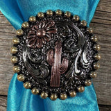 The Cactus Wild Rag Slide by Rodeo Drive features an antique silver concho with a cactus and flower design, gold bead border, and is displayed on turquoise fabric.