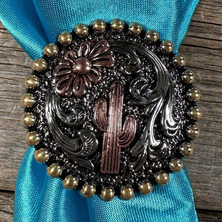 The Cactus Wild Rag Slide by Rodeo Drive features an antique silver concho with cactus and flower designs, accented with gold beads, styled on a turquoise wild rag atop a wooden surface.