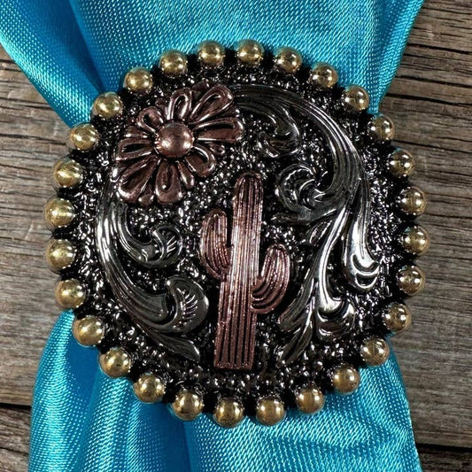 The Cactus Wild Rag Slide by Rodeo Drive features an antique silver concho with cactus and flower designs, accented with gold beads, styled on a turquoise wild rag atop a wooden surface.