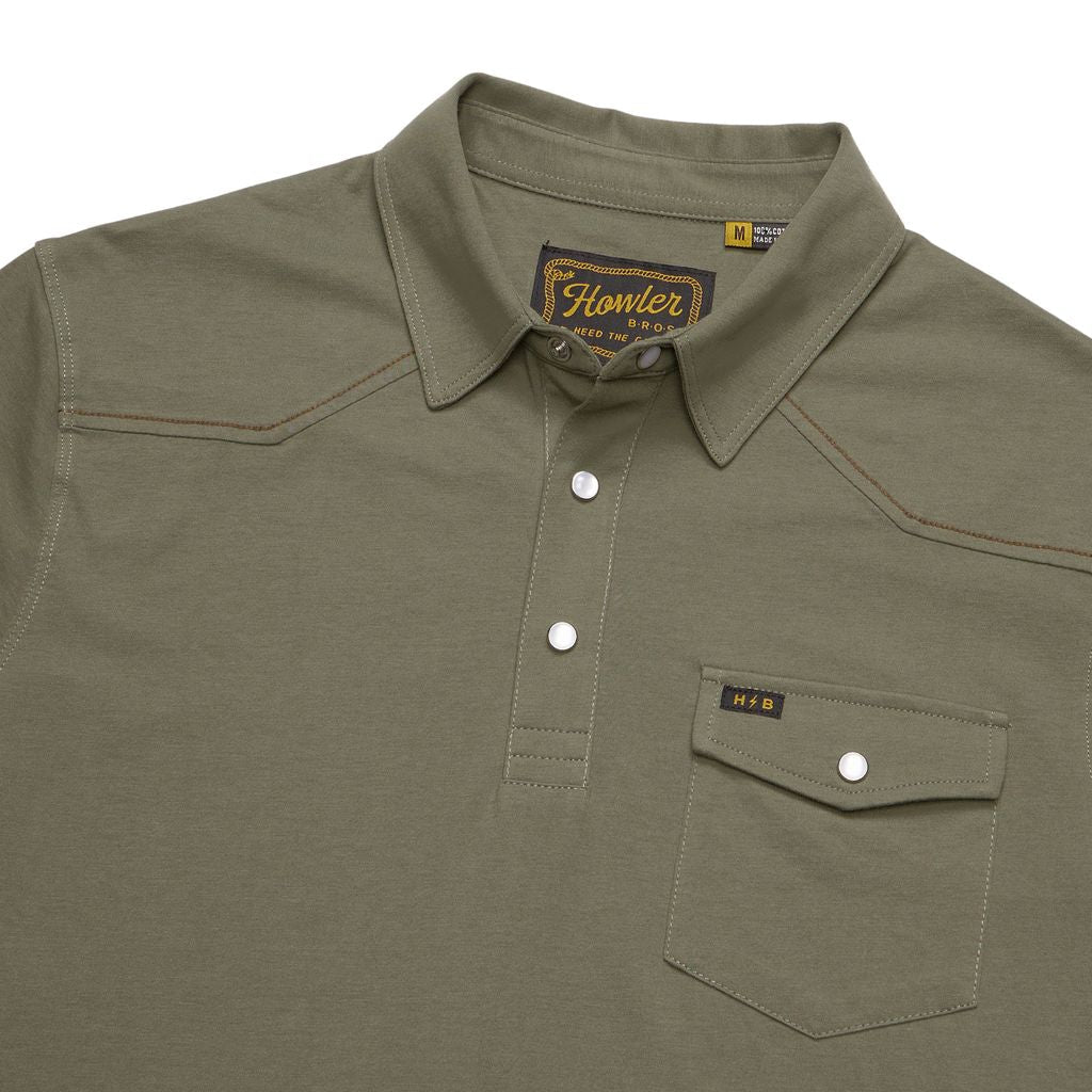 The Ranchero Polo by Howler Brothers features an olive green color with a collared neckline and three-button placket. It includes a chest pocket with a snap button, accented by brown stitching details. Subtle Western yoke styling enhances the design, while a stitched “Howler” label sits above the pocket.