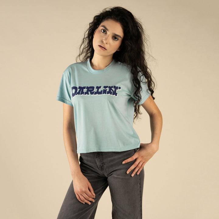 A woman with long curly hair wears the Sendero Provisions Co. "Call Me Darlin' Boxy Tee" in light blue with dark jeans, standing against a beige background—ideal for content about conversation tips or remembering names.
