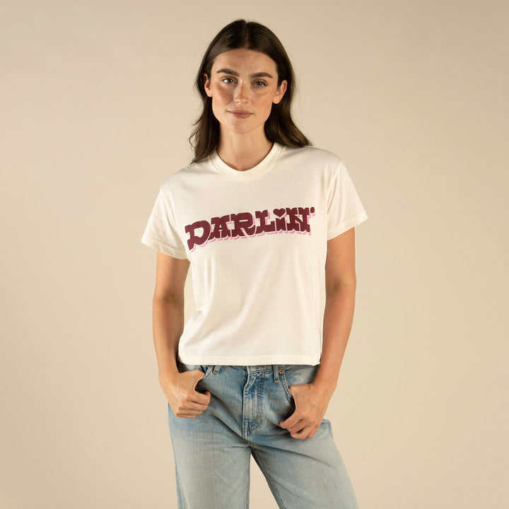 A woman poses against a beige background wearing the Sendero Provisions Co. Call me Darlin' Boxy Tee—an ultra-soft, white organic cotton shirt with “DARLIN’” in bold red letters—paired with light blue jeans.