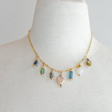 The Calypso Jewel Tone Charm Necklace by Jill Makes is a 16-inch gold chain adorned with seven assorted gemstone and bead pendants, showcased on a white bust against a plain background.