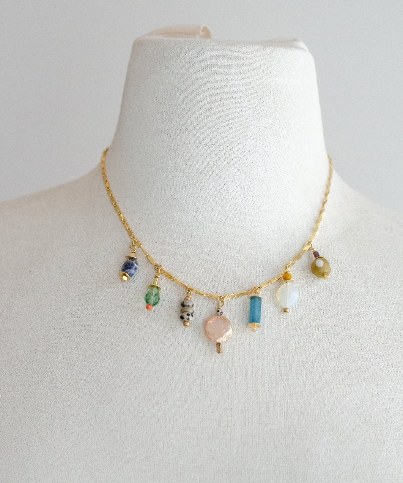 The Calypso Jewel Tone Charm Necklace by Jill Makes is a 16-inch gold chain adorned with seven assorted gemstone and bead pendants, showcased on a white bust against a plain background.