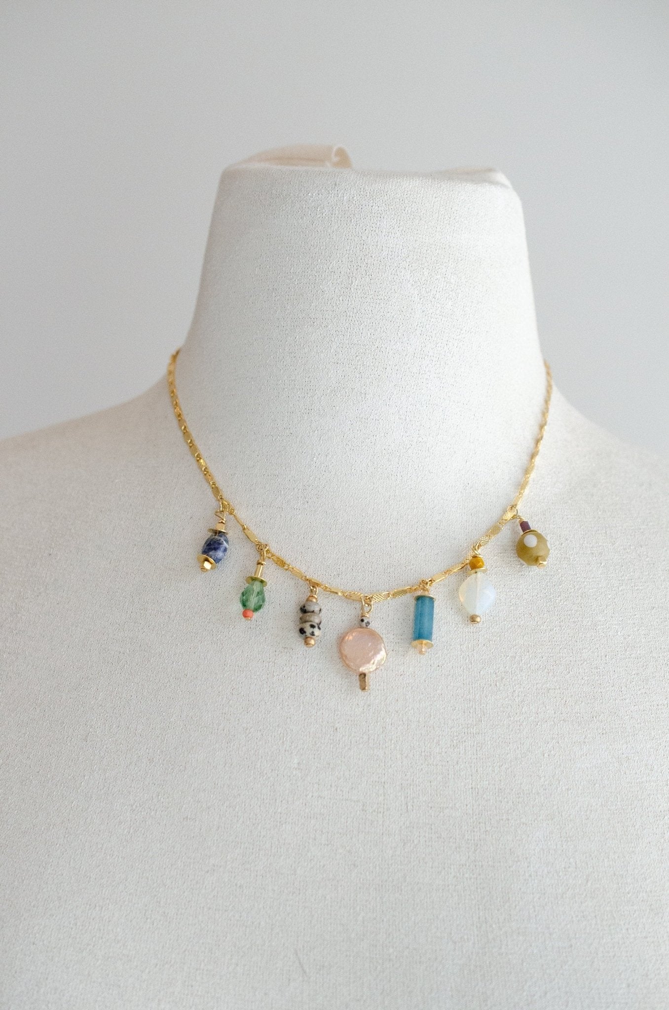 The Calypso Jewel Tone Charm Necklace by Jill Makes is a 16-inch gold chain adorned with seven assorted gemstone and bead pendants, showcased on a white bust against a plain background.