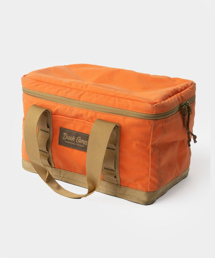 The Duck Camp Canvas Ammo Tote- Blaze is a rectangular orange canvas bag with tan handles and trim, zippered top, and front "Duck Camp" label—ideal as a durable tote for your outdoor essentials.