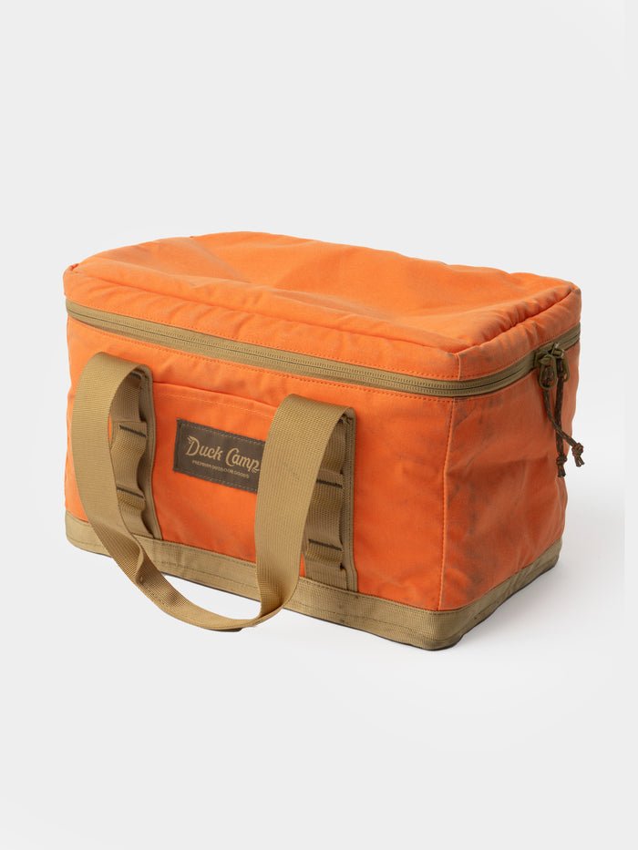 The Duck Camp Canvas Ammo Tote- Blaze is a rectangular orange canvas bag with tan handles and trim, zippered top, and front "Duck Camp" label—ideal as a durable tote for your outdoor essentials.