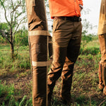 A person in an orange shirt and brown pants stands outdoors, holding the Duck Camp Canvas Gun Sleeve—a durable, brown waxed canvas case with beige straps—ideal for firearm protection, surrounded by grass and trees.