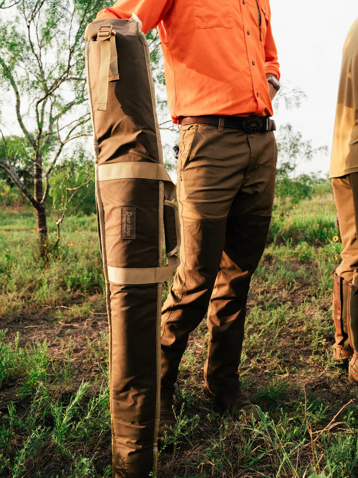 A person in an orange shirt and brown pants stands outdoors, holding the Duck Camp Canvas Gun Sleeve vertically on the ground.