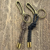 Two Tres Cuervos Canvas Ocho Keychains with braided canvas tassels—one brown, one black—each finished with brass tips and a figure 8 knot, are displayed on a wooden surface.